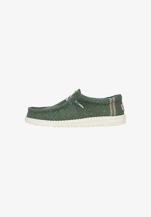 Zapato de tela verde sin cordones con suela de goma blanca, que presenta un sistema de lazado elástico y acentos estampados en los laterales. Superficie texturizada.