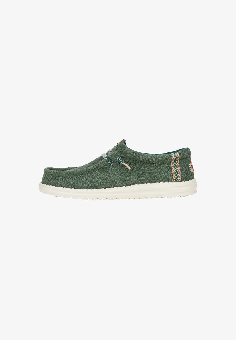 Scarpa slip-on in tessuto verde con suola in gomma bianca, dotata di lacci elastici e decorazioni con motivi sui lati. Superficie ruvida.