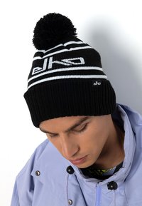 Zwart-witte gebreide beanie met een horizontaal gestreepd ontwerp, voorzien van een pompon bovenop en een merkpatch aan de voorkant.