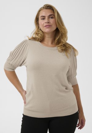 Frau mit blondem Haar, die einen beigefarbenen Kurzarm-Pullover mit plissierten Ärmeln und schwarze Hose trägt, steht mit einer Hand in der Hüfte.