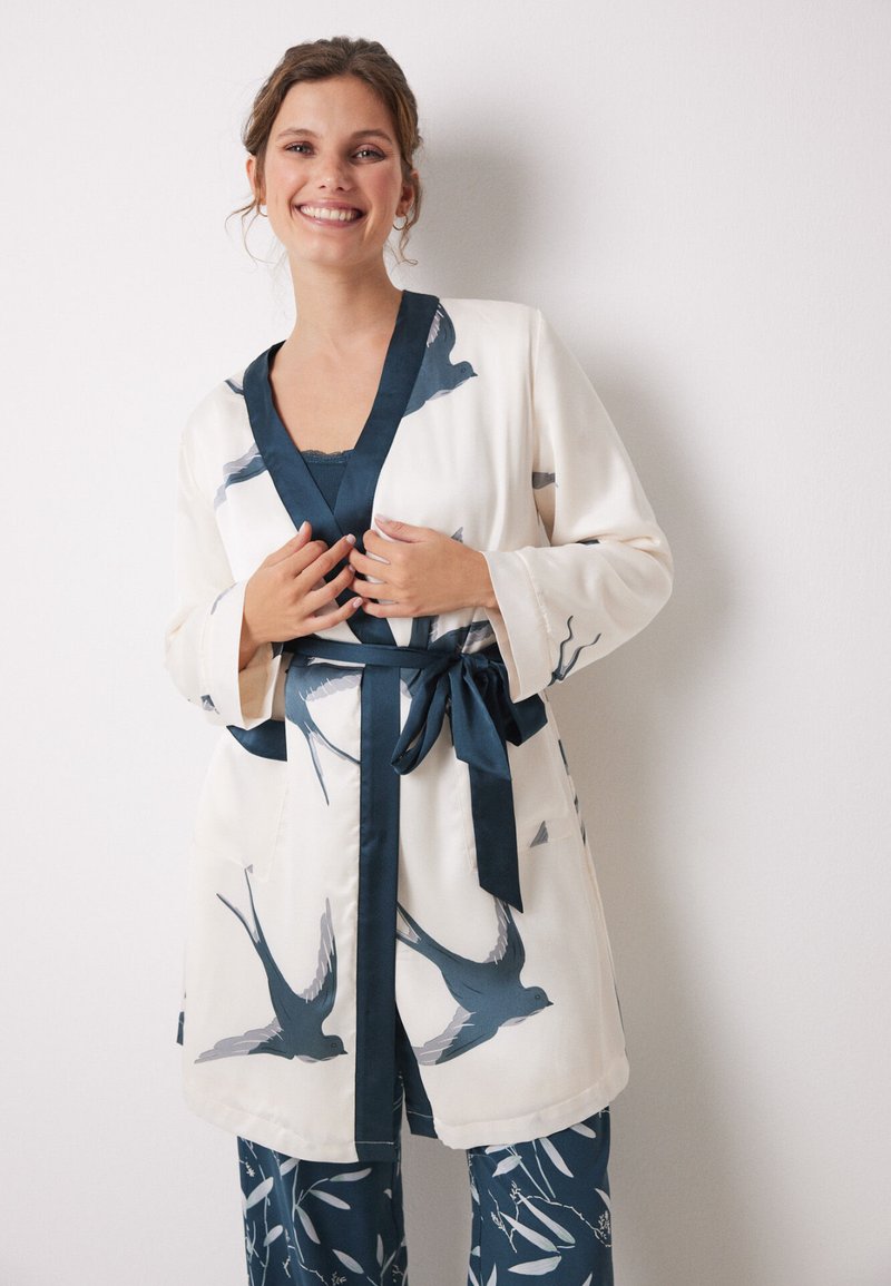 Accappatoio lungo in seta di colore bianco sporco con motivi di rondini blu e un bordo in satin blu scuro. Presenta una cintura in vita e maniche larghe; abbinato a pantaloni decorati.