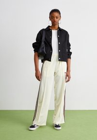 Schwarze Button-up-Jacke über einem weißen Hemd, cremefarbene Jogginghose mit kastanienbraunen Streifen und schwarze Sneakers; minimalistisches Design und lässige Passform.