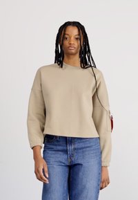 Beige cropped sweatshirt with long sleeves, side zipper detail, and ribbed cuffs. Worn with blue jeans. Smooth texture, casual design.
