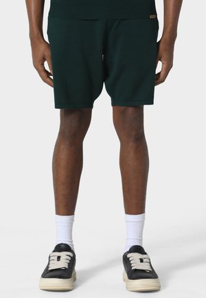 Mand iført mørkegrønne shorts, hvide crew-strømper og sorte sneakers med beige såler, stående mod en ensfarvet lys baggrund.