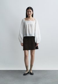 Femme portant une blouse blanche ample avec des détails en dentelle, une jupe marron foncé avec des poches, des chaussettes beiges et des ballerines noires, debout sur un sol en béton.