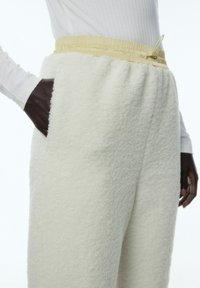 Weiße flauschige Fleece-Hosen mit einer strukturierten Oberfläche; verfügen über einen gelben elastischen Bund und seitliche Taschen für Funktionalität.