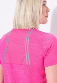 Donna bionda che indossa una maglietta sportiva rosa brillante con strisce riflettenti grigie, vista di spalle mentre gira leggermente la testa di lato.