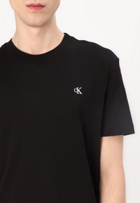 Persona que lleva una camiseta de manga corta negra con un pequeño logotipo blanco "cK" bordado en el lado izquierdo del pecho.