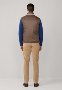 Gilet imbottito marrone sopra un maglione azzurro a trecce, abbinato a pantaloni in corduroy color marrone chiaro e scarpe scure, visto da dietro.