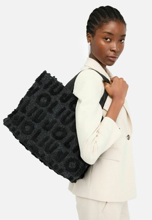 Black tote bag with textured fabric, featuring embossed letters, sturdy handles, and a square shape, carried by a woman in a light suit.