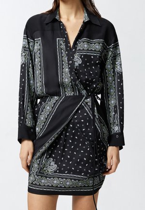 Femme portant une robe portefeuille noire à manches longues avec des motifs cachemire blancs et verts ainsi que des motifs géométriques.