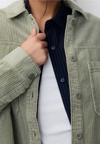 Chaqueta de camisa de pana verde oliva con una superficie texturizada, que presenta cierres de botón y un bolsillo. Debajo, se puede ver una camisa de rayas azul marino.