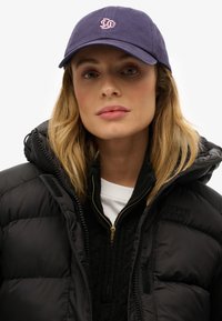 Mujer con una gorra de béisbol azul marino y una chaqueta acolchada negra cerrada sobre un suéter de punto negro con una camisa blanca debajo.