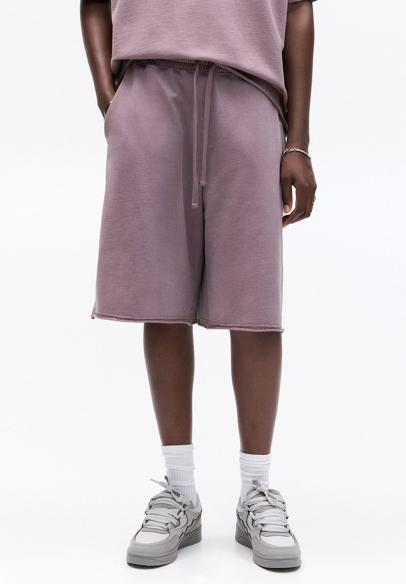 Personne portant un short mauve jusqu'aux genoux avec cordon, des chaussettes blanches montantes, des baskets grises et un haut mauve assorti, debout les mains le long du corps.