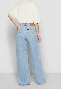 Personne portant un jean taille haute à jambes larges de couleur bleu clair et un haut blanc à manches courtes, avec une main dans la poche arrière, debout devant un fond uni.