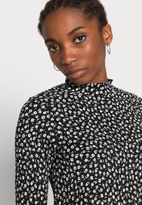 Camisola preta de manga longa com padrão floral branco, gola alta, design justo, tecido texturizado e um brilho subtil.