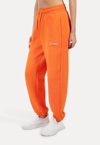 Orangene Jogginghose mit einem Kordelzugbund und gerippten Bündchen. Mit gesticktem Logo an der Seite. Hergestellt aus weichem, dickem Stoff.