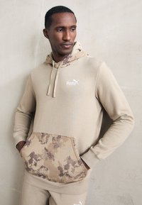 Sudadera beige con un bolsillo y forro de capucha de patrón de camuflaje. Presenta un logo blanco en el pecho y puños acanalados. Tejido suave y texturizado.