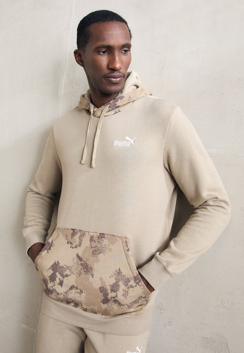 Sudadera beige con un bolsillo y forro de capucha de patrón de camuflaje. Presenta un logo blanco en el pecho y puños acanalados. Tejido suave y texturizado.