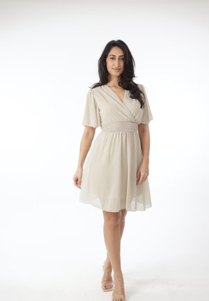 Mujer con cabello largo y oscuro, vestida con un vestido beige hasta la rodilla, con mangas cortas y sueltas y cintura fruncida, de pie contra un fondo blanco.