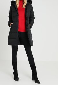 Piumino nero con cappuccio in pelliccia sintetica, maniche lunghe e chiusura con cerniera. Indossato sopra una maglia rossa, abbinato a pantaloni skinny neri e stivali alti.