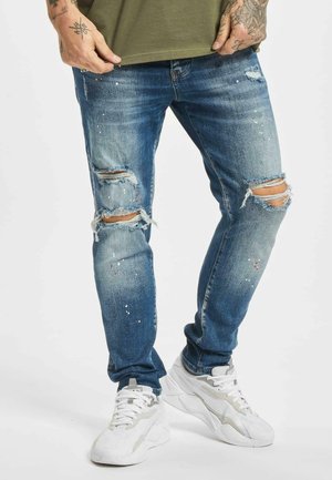 Jeans de mezclilla azul rasgados con detalles desgastados y salpicaduras. Llevados con zapatillas blancas, mostrando un ajuste relajado y pierna ceñida.