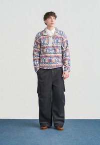 Noor mees seisab käed taskutes, kannab värvilise mustriga pusa, laia musta taskupükse ja pruune kingi.