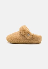 Tan kleurige fluffy instap pantoffels met een zachte band en een ronde embleem, tegen een plain witte achtergrond.