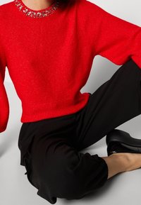 Maglione rosso a maglia con scollatura decorata da gioielli, abbinato a pantaloni neri. Il maglione ha una vestibilità ampia e polsini a coste, mentre i pantaloni hanno una silhouette morbida.