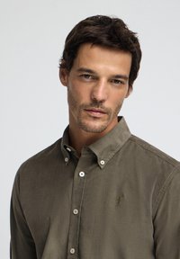 Olivgrön skjorta med knappar, gjord av mjukt tyg; har en liten broderad logotyp på bröstet och klassisk krage.