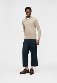 Άνδρας που στέκεται φορώντας μπεζ πουλόβερ με φερμουάρ μέχρι τη μέση, σκούρο παντελόνι με φαρδιά, κομμένα γυαλιστερά τζιν, μπεζ κάλτσες και καφέ δερμάτινα παπούτσια σε λευκό φόντο.