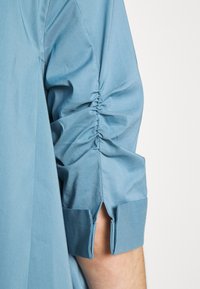 Manche de chemise bleue avec tissu froncé le long du bras et une petite fente au-dessus du poignet laissant apparaître la peau en dessous.