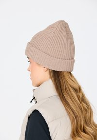 Beige ribbade stickade mössa med vikkrage, konisk topp, bärs av en person med långt hår. Bakgrunden är helt vit.