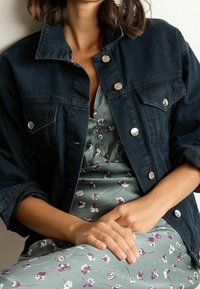 Chaqueta de mezclilla oscura con botones de presión plateados y dos bolsillos en el pecho, llevada sobre un vestido verde con estampado floral. Textura suave y lisa.