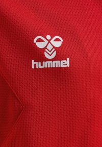 Białe logo marki Hummel i tekst na czerwonym, teksturowanym materiale sportowym z ukośnym szwem po lewej stronie.