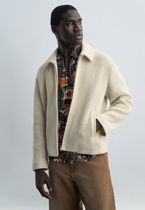 Vīrietis, tērpies krēmkrāsas džemperī pār melnu krekla ar oranžām un baltām ziedu rakstiem, valkā brūnas bikses un skatās sānis.