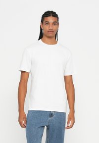 Young man with braided hair wearing a plain white short-sleeve t-shirt and blue jeans, standing against a white background.