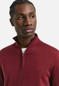 Bordeauxfarbener Zip-Pullover aus weichem Stoff, mit einer strukturierten Oberfläche und einem dezenten Logo auf der Brust.