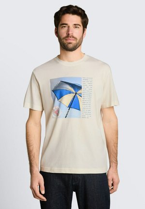 Mężczyzna ubrany w beżowy t-shirt z niebiesko-beżową grafiką parasola plażowego i napisem, w połączeniu z ciemnymi dżinsami, stoi na jednolitym tle.