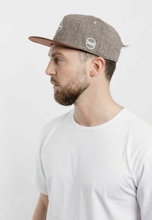 Smeđi, teksturirani snapback šešir s ravnim obodom, s bijelim logotipom i detaljima; nosi se uz običnu bijelu majicu.