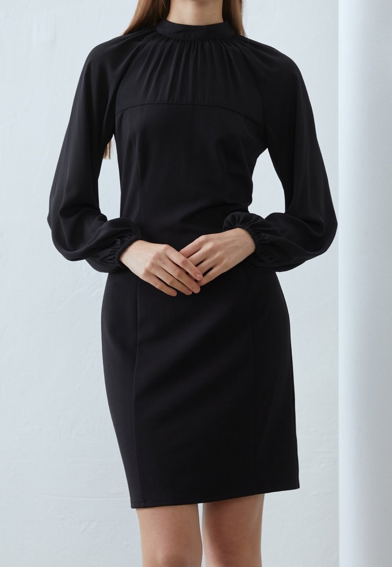 Femme portant une robe noire à manches longues avec poignets froncés et col montant, debout les mains doucement jointes devant elle.