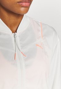 Valkoinen kevyt bomber-takki, jossa on vetoketjukiinnitys, pinkit yksityiskohdat ja pieni Puma-logo rintakehässä. Sileä pinta, urheilullinen muotoilu.