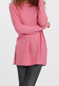 Pitkä vaaleanpunainen neulepaita, joka on valmistettu pehmeästä neulemateriaalista. Paidassa on väljä leikkaus, sivuhalkiot ja hieman kaareva helma, joka on yhdistetty tummiin farkkuihin.