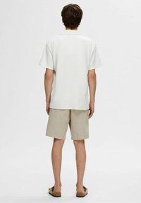 Young man seen from behind wearing a white short-sleeve shirt, beige shorts, and sandals against a plain light background.