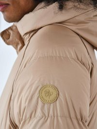 Beige pufferjakke med en markant tekstureret stof. Har en hætte, rund krave og et guld-logo patch på venstre skulder.