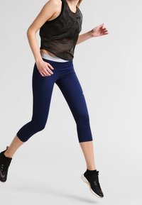 Top sin mangas de color gris oscuro con el logo "NIKE", combinado con leggins de capri de color azul marino. El modelo lleva zapatillas deportivas negras y calcetines hasta el tobillo.