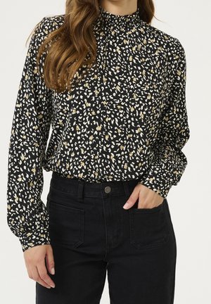 Femme portant un chemisier noir à manches longues avec des taches abstraites blanches et dorées, rentrée dans un jean noir taille haute.