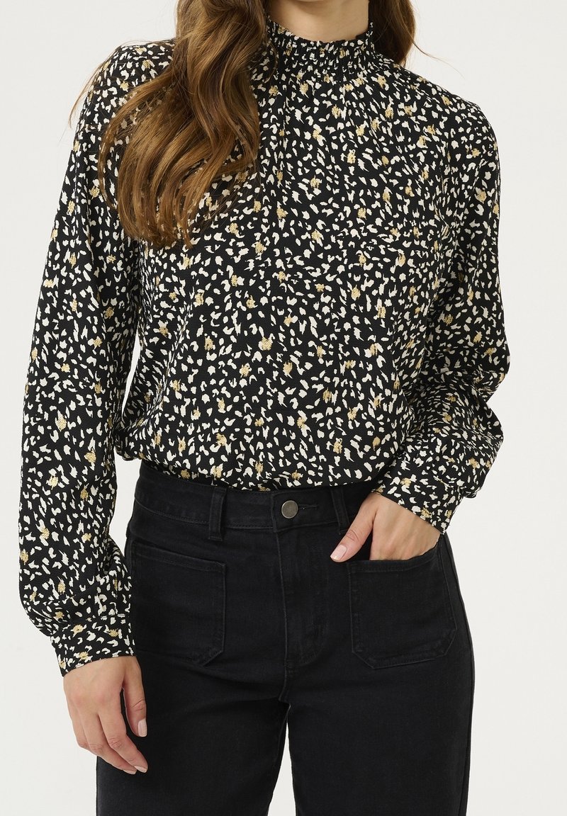 Femme portant un chemisier noir à manches longues avec des taches abstraites blanches et dorées, rentrée dans un jean noir taille haute.