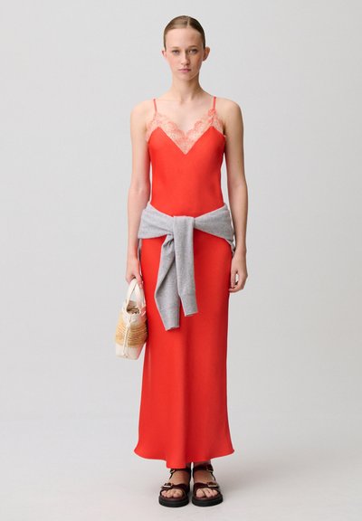 Donna in piedi che indossa un abito sottoveste rosso con rifiniture in pizzo, maglione grigio annodato in vita, tiene una borsa beige e indossa sandali neri su sfondo neutro.