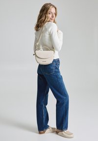Beige strukturierte Handtasche mit einem runden oberen Teil, verstellbarem Trageriemen und strukturierter Oberfläche. Das Model trägt einen weißen Pullover und blue jeans.